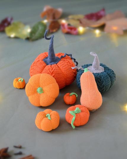 Ensemble de citrouilles et courges crochetées de tailles et de couleurs variées (orange, vert foncé et orange doux), posées sur un fond gris/vert avec quelques feuilles d'automne et une guirlande lumineuse floues en arrière-plan.