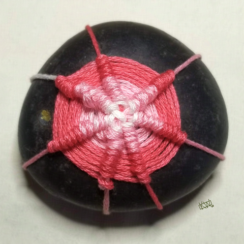 Photo of a round black stone wrapped in pink embroidery thread woven in a spider web stitch.
