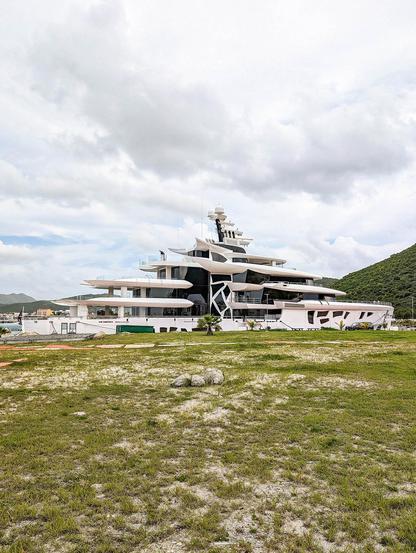 It's an overcast day with a few patches of blue sky, but it's hot and humid here. We're looking across a stretch of grass, patchy with dry spots in places. In the distance there are some small hills rising but they're mostly obscured by the superyacht that's moored in front with its upper decks visible but most of its hull below the level of the ground we're gazing past. The yacht structure is broadly white in colour but where it has large glass windows they're tinted almost to black, probably for privacy. The effect is almost as if a large block of smoky quartz were broken up by slices of white decking arranged almost organically, slightly offset vertically. A central column seems to show a central atrium on the yacht of four decks with window shapes as if rectangles had been stretched and warped and twisted. A very strange-looking ship.