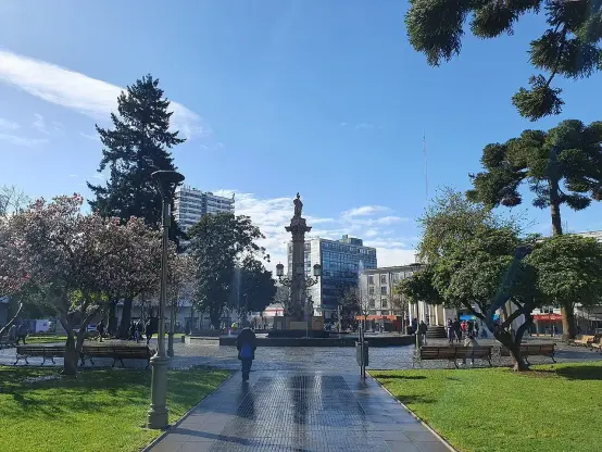 The image depicts a public park or plaza with a central monument. There are trees, benches, and some flowering plants surrounding the area. The sky is clear with a few clouds, indicating a bright day. People are visible in the distance, enjoying the space. The layout suggests it's likely a place for relaxation and leisure.

Image Credits: Wikimedia / TomasVial / CC BY-SA 4.0