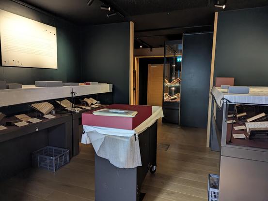 A darkened exhibition room with dark green walls, exhibition cases on either side and ahead covered in white protection. Trolley at centre of space with large box on top awaiting return of manuscript.