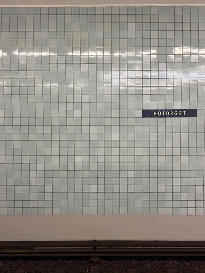 Photo of a wall in a metro station. The name of the metro station is visible on a dark blue sign with white letters saying ”Hötorget”. One of the more central stations in Stockholm. The wall is filled with tiles in a light bluish tone. There is a variation in level of blue. Tiles measure about 10x10 cm or 15x15 cm. Reflection of platform lighting can be seen in the upper part of the tile filled wall.