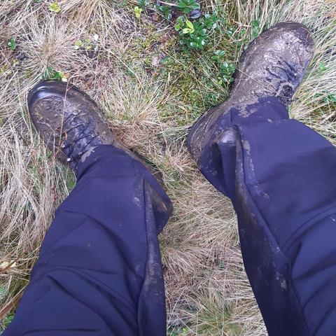 Photo of lower legs and feet of a person wearing black pants and hiking boots  standing in the grass. The boots are covered in fresh, wet mud, and the pant legs are also dirty.