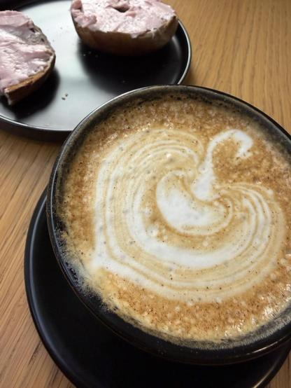 A closeup of a latte, with a pumpkin drawn into the foam. There's a bagel with strawberry cream cheese in the background.