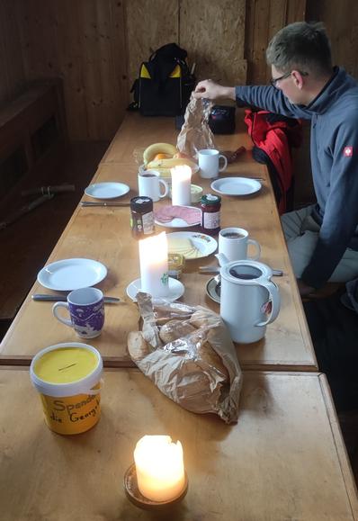 Gedeckter Frühstückstisch mit Tellern, Tassen, Kannen. Einer sitzt schon dran. 3 Kerzen leuchten.