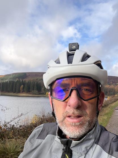 A man with gray facial hair is wearing a white cycling helmet and sunglasses. He is standing near a lake with a scenic view of hills and trees in the background. The sky is partly cloudy.
