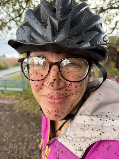 A person wearing glasses and a black helmet smiles, with a face covered in dirt splatters. They are dressed in a pink jacket and are outdoors, likely after riding a bicycle on a dirt path. Trees and a bench are visible in the background