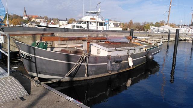 Kleiner Tradisegler im winterlichen Yachthafen. Lack ist stumpf, ein langer Bart unter Wasser, rig ist abgetakelt und die Masten liegen an Deck