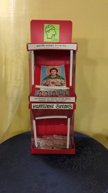 Sticker totem con adesivi in omaggio. Sopra il santino di Luigi Mangione.