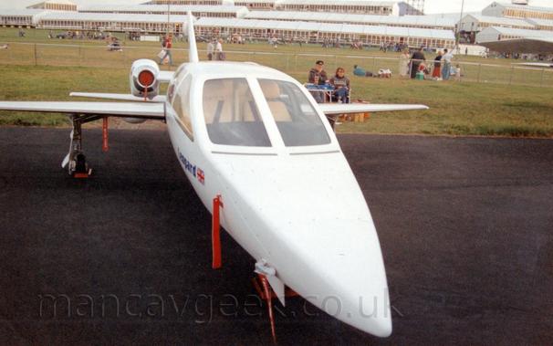 Head-on view of a small, twin engined, 4 seat BizJet, parked on black tarmac at an air show.
The plane is rather angular, with a pair of small jet engines attached to the sides of the rear fuselage.
The cockpit  has large windows, with a pair of pretty comfortable-looking seats visible through the front windows.
A man with a black cap and a woman with reddish brown hair are sitting at a collapsable table behind the plane.
People are walking past fencing in the distance, with large temporary buildings rising up the side of a hill in the distance.