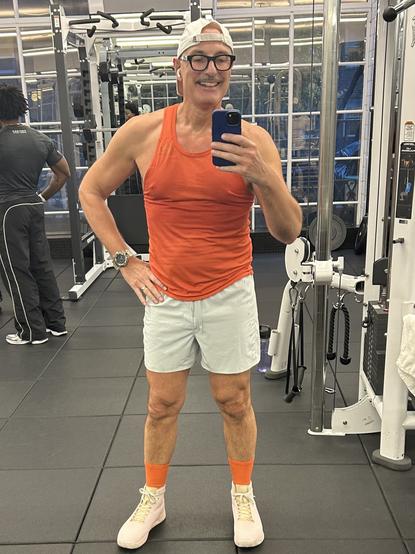 A muscular man (me?) is posing in a gym, wearing an orange tank top, light gray shorts, and pink sneakers with orange socks. He is holding a phone for a selfie. In the background, gym equipment and another person can be seen.