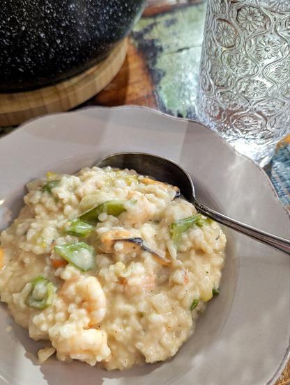 Tiefer, weißer Porzellanteller, darin eine cremige, helle Reispampe mit Meeresfrüchten und grünen Stücken von Kaiserschoten. Ein Löffel liegt im Teller. Daneben auf dem Tisch ein großes Wasserglas mit geprägtem floralem Muster, ein schwarzer Topf auf einem Holzbrett.