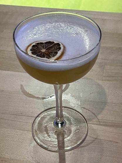 A coupe-style cocktail glass sits on a wooden surface. It's filled about halfway with an opaque yellowish liquid. A layer of white foam rests on top, and in the foam floats a circle of candied blood orange: nearly white pith forming a wagon-wheel pattern against the deep red of the flesh of the orange.