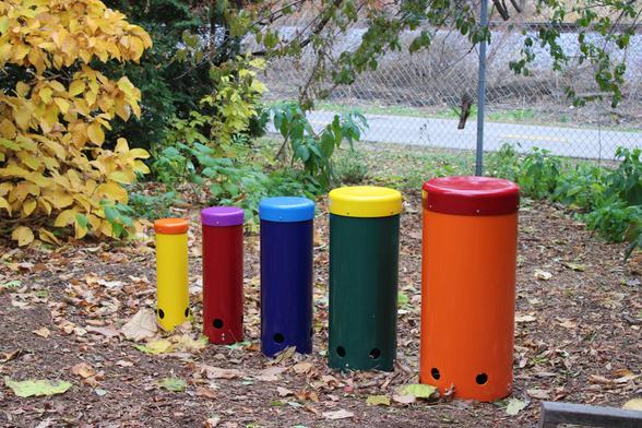 Brightly painted metal cylinders of different heights with some dried fallen leaves in front of a bush with yellow and brown leaves and a metal chain link fence with an asphalt bike path on the far side.