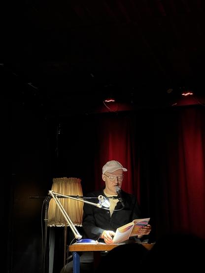 A person wearing a white cap and glasses is sitting at a table with a microphone, reading from a book. The setting features red lighting, a lamp with a shade, and a dark background, indicating an intimate performance or reading event.