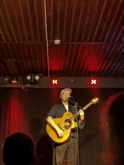 A musician is performing on stage with an acoustic guitar. He is wearing glasses and a cap, and the background features red lighting against a black curtain. The venue has a wooden ceiling, and part of the audience is visible in the foreground.