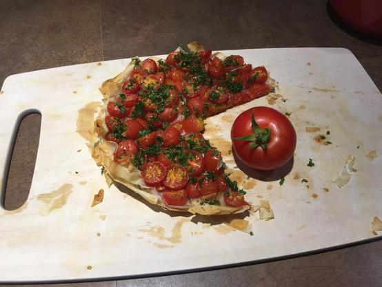 A round tart, packed with cherry tomatoes, on a cutting board, with a quarter of it missing. There is a larger red tomato in the empty quarter, making it look like pacman eating a tomato.