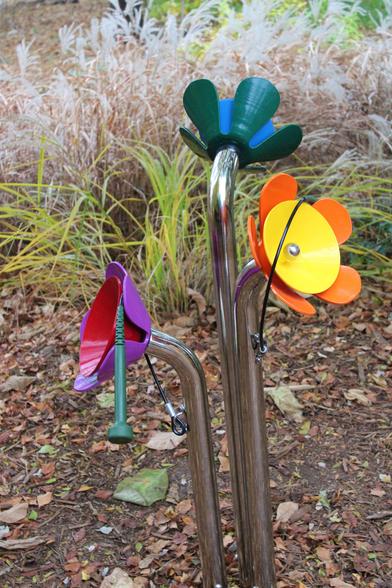 Chrome pipe stems in the bare ground attached to brightly colored two-layered metal dishes with the outer dish shaped to resemble flower petals. A green striking tool on a black plastic cord is dangling from the purple and red dishes on the left side. Decorative grasses are growing in the background.