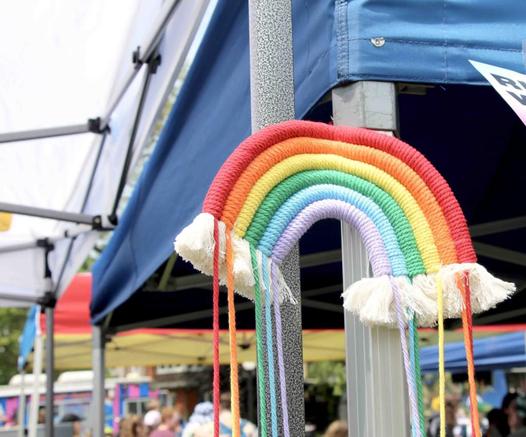 Photo of a woven fabric rainbow, with white tassels trimming each end like clouds, hanging from a booth during Tri Pride in Kitchener, Ontario.