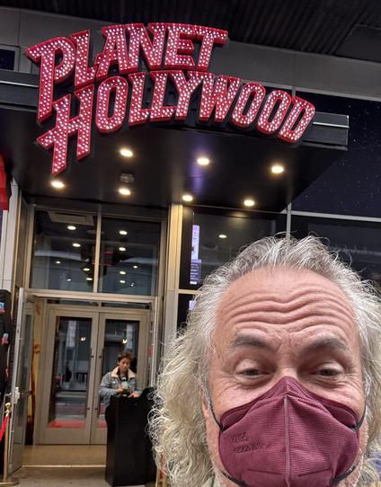 Selfie of respirator-wearing man on sidewalk , over his shoulder is the entrance to Planet Hollywood.