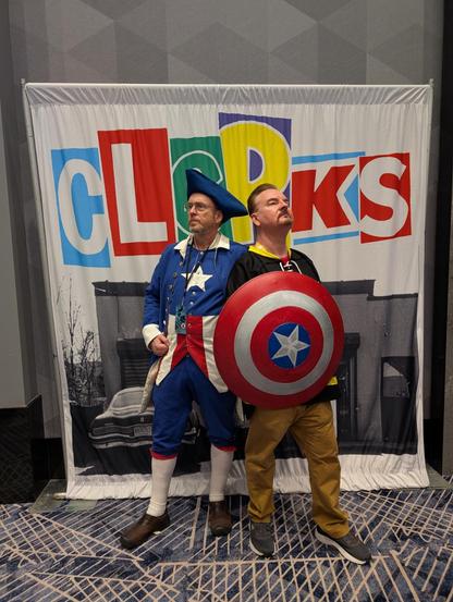 A man cosplaying as Colonial America standing next to Brian O'Halloran in front of a Clerks banner. Brian is holding Captain America's shield