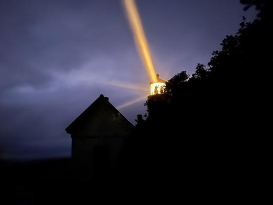 Orange beams of light shine out of the Heceta Head Lighthouse against a purplish cloudy sky. An outbuilding and forest are silhouetted in the foreground. Photo by Robert Emond.