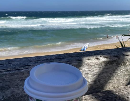 Takeaway coffee up on a weathered wooden bench overlooking the sand and waves at Bar Beach. There are two white and blue lifeguard rescue boards (surfboard shaped) resting on the sand. Blue skies, sunshine and lots of white from breaking waves in green-blue water.