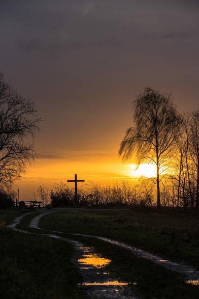 Man sieht ein Gipfelkreuz das rechts und links von Bäumen umgeben ist. Links unten steht Bänke die zur Rast einladen. Es führt ein geschwungener Weg zu dem Aussichtspunkt, in kleinen Pfützen spiegelt sich das Licht der aufgehende Sonne.
Die scheint durch eine Lücke, im ansonsten stark bewölkten Himmel.