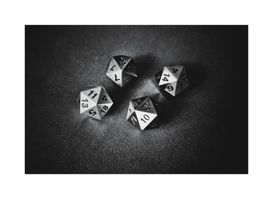 A high-angle, close-up, black and white photograph of four metallic twenty-sided dice (d20) resting on a dark, velvety surface. The dice are casually grouped, with various numbers visible on their top faces. The lighting creates strong highlights and shadows, emphasizing the sharp edges and numbering of the dice.