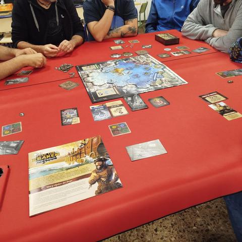 Ocho personas juegan en torno a dos mesas juntas sobre las que se despliega el tablero y cartas del juego Feed the Kraken.