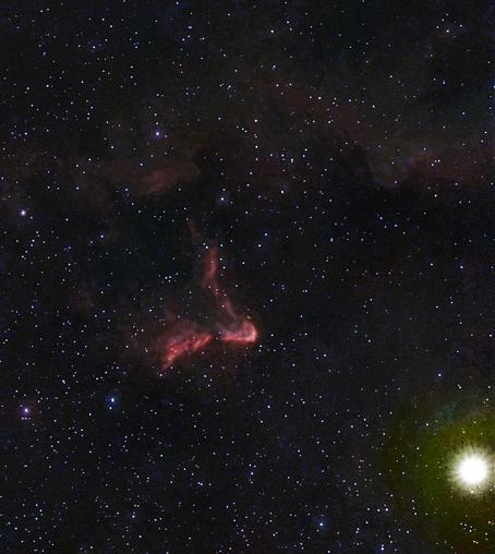 Rozległy widok przestrzeni kosmicznej z licznymi gwiazdami na ciemnym tle, z widoczną jasną gwiazdą po prawej stronie i czerwonawą mgławicą tworzącą charakterystyczny kształt w środku po lewej stronie.