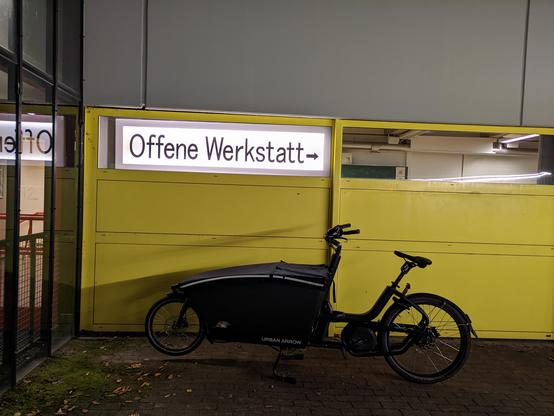 Gelbe Wand mit schmalen Fenstern im oberen Bereich. In einem Fenster hängt ein beleuchtetes Schild "Offene Werkstatt ->"