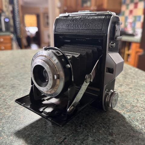An Ensign Selfix 16-20 sits on a kitchen work top. The Selfix is a folding camera with a silver lens assembly attached to a leather bellows that when the front on the camera is closed the whole assembly folds up.