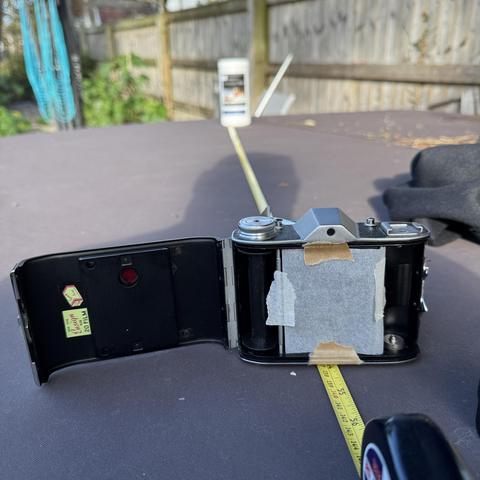 An Ensign Selfix sits on a brown box. The back is open and the film area  covered by a sheet of grease proof paper. Under the camera is a yellow tape measure and the far end of the box (4.5” / 1.37m away) is a white plastic container.