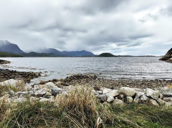 Rocky shoreline foregrounds a body of water with small islands and mountainous landscape under a cloudy sky.