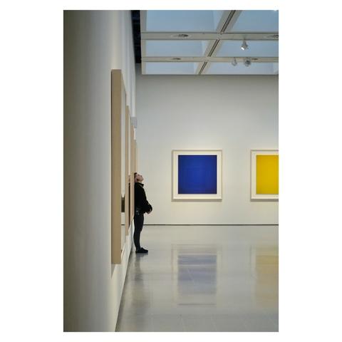 This is a portrait format colour image of the inside of a modern art gallery. Taken looking along a wall, a security attendant is viewed from the side in mid-frame with their back against the wall. They are looking up to the flat glass ceiling. To the back are large colourful square images on display: one blue and one yellow. They are reflected in the shiny white gallery floor. Taken in London with a Sony RX100.
