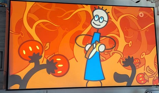 Foto der Projektorleinwand mit einem Ausschnitt einer Tom und sein Erdbeermarmeladebrot mit Honig folge.

Tom - eine comic Figur mit blauem Shirt und Brille steht in einem dunklen Wald, der durch gerade leicht beleuchtet und damit rötlich wirkt. Im Vordergrund sind Erdbeeren an den Pflanzen zu sehen. Tom hält ein Brot mit Honig in der Hand.