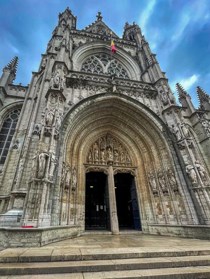 Exterior of Église Notre–Dame au Sablon in Brussels, seen from below—Gothic façade with statues and open doors beneath a cloudy blue sky.