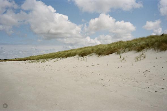 Bright, wide sand dunes with sparse beach grass under a loosely cloudy blue sky; peaceful, unspoilt coastal landscape.