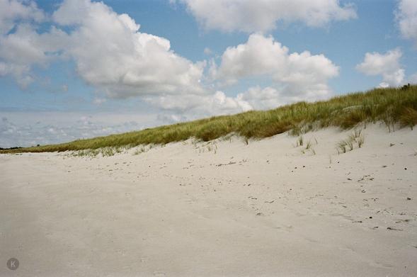 Bright, wide sand dunes with sparse beach grass under a loosely cloudy blue sky; peaceful, unspoilt coastal landscape.