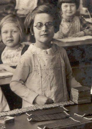 A photo of a young girl in glasses. She is standing behind a school bench. Other girls are visible behind her.