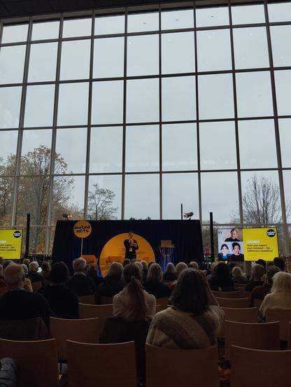 A wide shot of an indoor event, likely a lecture or presentation, taking place in a large room with floor-to-ceiling windows. A person is standing on a stage, facing an audience seated in rows of chairs. The stage has a dark backdrop with a yellow circular design featuring the text “malmö” in a stylized font. On either side of the stage are vertical banners with images of people and additional text. The windows behind the stage create a grid-like pattern, and the room appears to be well-lit. 

The banners on the left side have text that reads “sem vapen” and “malmö” in a stylized font. The banners on the right side have text that reads “ord som” and “malmö” in a stylized font. The audience is mostly facing forward, and the seats are filled with a variety of people. The color scheme is mostly neutral, with browns, yellows, and blacks dominating the scene.

Provided by @altbot, generated privately and locally using Gemma3:27b

🌱 Energy used: 0.139 Wh