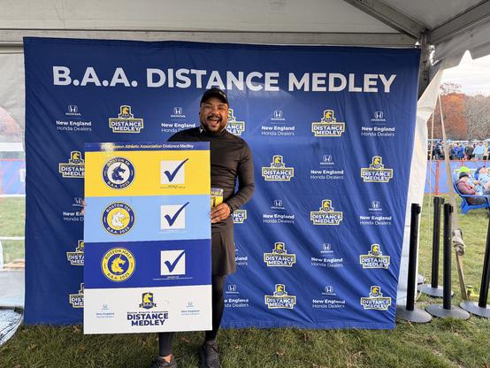 Photo of me standing in front of a blue and white backdrop that says “B.A.A. Distance Medley”

I’m holding a poster board with 3 check marks next to the three races of the medley: 5K, 10K, Half Marathon