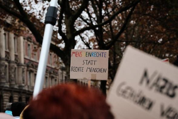 Eng gedrängte Menschen vor einem Gebäude mit Säulen. Auf einem Pappschild ist "Menschenrechte statt rechte Menschen" zu lesen"