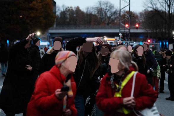 Mehrere Menschen in dunkler Kleidung stehen in einer Reihe und schwenken Mobiltelefone mit Licht. Davor stehen zwei weitere Personen mit roten Jacken einander zugewandt, als ob sie gemeinsam singen. Die Gesichter sind unkenntlich gemacht.