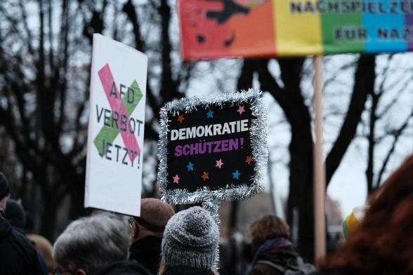 Menschen stehen eng gedrängt unter kahlen Bäumen. Über den Köpfen ist ein Schild "AfD-Verbot jetzt" und ein mit Lametta umwickeltes Schild mit der Aufschrift "Demokratie schützen" zu sehen