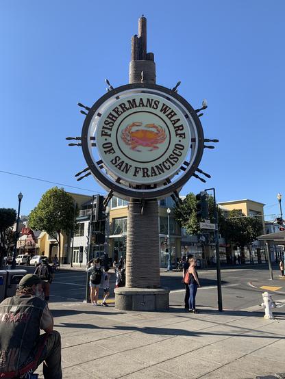 Circular sign reading "Fisherman's Wharf of San Francisco" mounted on top of a tall, textured pillar in an urban street setting. Below the sign, a person seated wearing a dark jacket and jeans captures the scene on camera. Other pedestrians are visible in the background near street corners and storefronts. Clear skies and sunlight illuminate the scene.