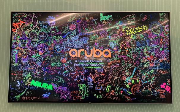 Colorful graffiti-style artwork covers a large, illuminated board, prominently featuring the word "Aruba" under the tagline "a Hewlett Packard Enterprise company." The artwork is dense with vibrant, neon drawings and text in various colors, including sketches of faces, words, and abstract designs all interconnected, creating a lively and chaotic visual impression against a dark background.