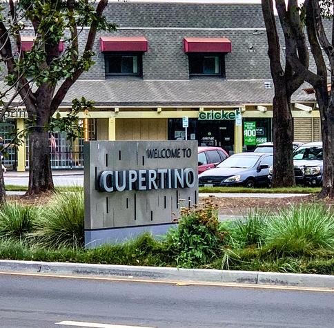 Gray sign reading "WELCOME TO CUPERTINO" in bold cut-out letters, positioned in front of a landscaped area along a street. In the background, a two-story commercial building with a green Cricket Wireless store and parked cars are visible.