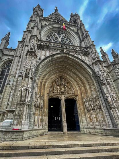 A close-up view of a grand, intricately detailed Gothic church entrance, featuring a large archway with sculptures and a flag above. The facade is adorned with stone carvings, and the stairway leading up to the doors is visible.